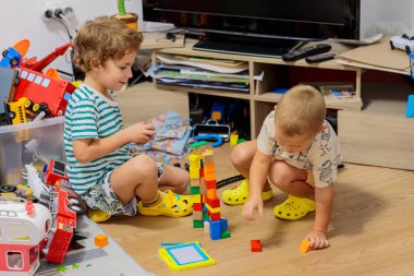 Two little boys playing on the floor with colorful construction blocks in a messy living room, surrounded by toys and a TV stand