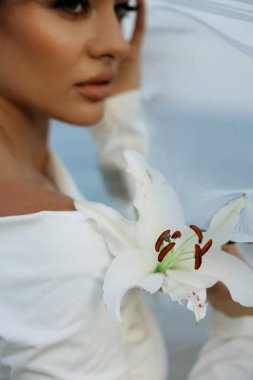 Beyaz elbiseli gelin elinde beyaz Lilly ile duvağıyla denizde poz veriyor. 