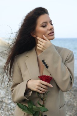 Kumsalda bej takım elbiseli güzel esmer kadın elinde kırmızı bir gül, yüzüne dokunuyor. 