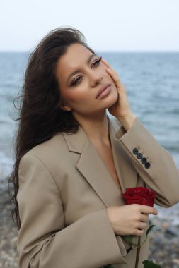 Kumsalda bej takım elbiseli güzel esmer kadın elinde kırmızı bir gül, yüzüne dokunuyor. 
