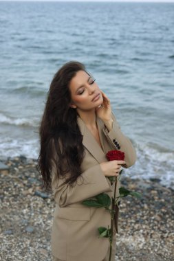 Kumsalda bej takım elbiseli güzel esmer kadın elinde kırmızı bir gül, yüzüne dokunuyor. 