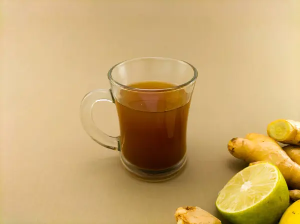 Şeffaf cam bardak sıcak zencefil suyuyla dolu, açık kahverengi arka planda duruyor.