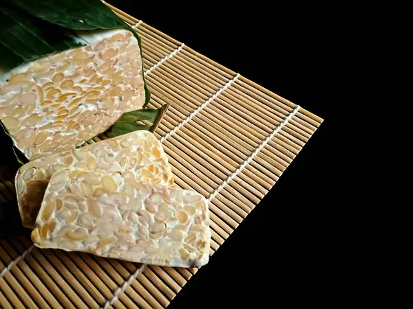 Geleneksel bir Endonezya mayalanmış soya fasulyesi keki olan Tempeh 'e yakın plan, kısmen muz yaprağına sarılmış. Siyah arka planda izole edilmiş, sağlıklı bitkisel gıdalar sergiliyor..