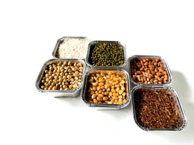 Temiz beyaz bir yüzey üzerinde farklı alüminyum folyo kaplarında sıralanmış çeşitli ham tahıl ve baklagillerin üst görünümü. Sağlıklı beslenme, gıda çeşitliliği, vejetaryen aşçılık, beslenme ve sürdürülebilir yaşam için ideal temalar.