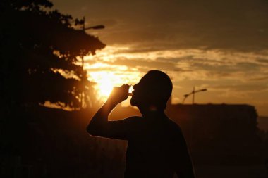 Gün batımında kadehten yudumlayan şık bir adamın silueti. Güneş ışığı, gökyüzünü turuncu ve sarıya boyar. Nostaljik ve sakin bir atmosfer yaratır. Sahne huzurlu bir günün sonunu yakalar..