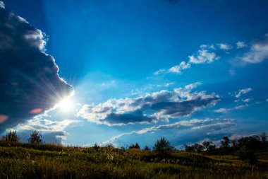 Düz bir düzlükte parlayan bulutlar ve güneş ışınlarıyla dolu mavi bir yaz gökyüzü..