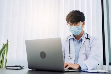 Doktor ofiste oturmuş, maske takarken dizüstü bilgisayarla uğraşıyordu..
