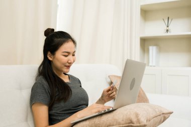 Asyalı bir kadın evde tatildeyken kanepede otururken online alışveriş için kredi kartı tutuyor..