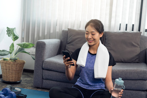 Asian woman playing smartphone at home during break from yoga exercise and drinking fresh water.