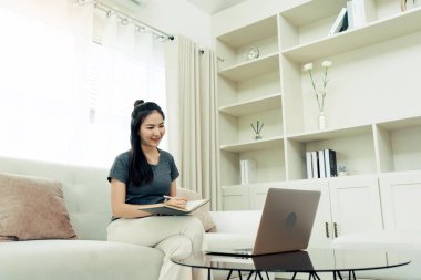 Asyalı bir kadın çalışırken ya da evde ders çalışırken bilgisayar ekranından notlar alırken kanepeye oturur..