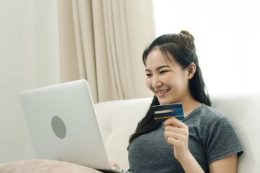 Asyalı bir kadın evde tatildeyken kanepede otururken online alışveriş için kredi kartı tutuyor..
