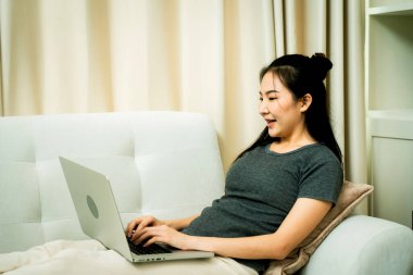 Asyalı kadın evde çalışmak için dizüstü bilgisayar kullanarak kanepeye oturuyor..