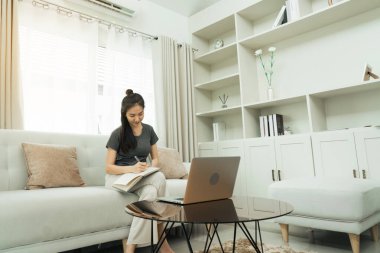 Asyalı bir kadın çalışırken ya da evde ders çalışırken bilgisayar ekranından notlar alırken kanepeye oturur..