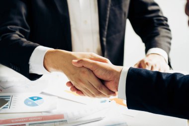 Girişimcilerin işbirliği anlaşması modern bir ofiste el sıkışıyor ve masadaki finansal kağıt grafiği.