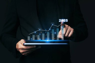 Tablet tutan ve 2025 yılındaki eğilimli piyasa büyümesi grafiğinde şirket finansmanı hakkındaki metni işaret eden işadamı, 2025 yılında.