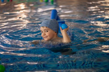 Küçük çocuk havuzda makarnayla yüzmeyi ve kapalı havuzda mavi topla oynamayı öğreniyor.