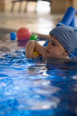 Canlı mavi bir havuzda renkli toplarla çevrili yüzme cihazıyla yüzme pratiği yapan küçük bir çocuk, neşeli anların tadını çıkarıyor.