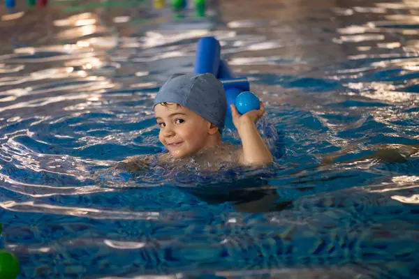 Küçük çocuk havuzda makarnayla yüzmeyi ve kapalı havuzda mavi topla oynamayı öğreniyor.