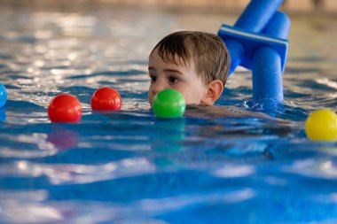 Küçük bir çocuk yüzme aleti kullanarak yüzmeyi öğreniyor ve kapalı havuzda renkli toplarla oynuyor.