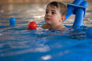 Küçük çocuk yüzme cihazıyla yüzmeyi öğreniyor, topla oynuyor, bilardoda eğleniyor.