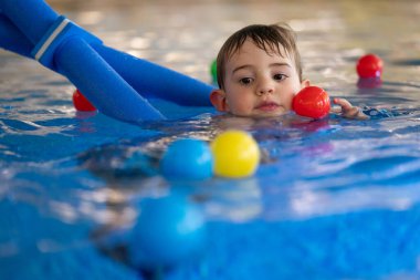 Küçük çocuk havuzda yüzme cihazının yardımıyla yüzmeyi öğrenirken renkli toplarla oynamaktan hoşlanıyor.