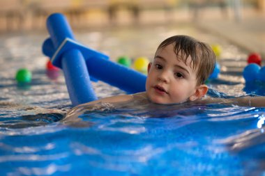 Küçük çocuk, kapalı havuzda yüzme aygıtı kullanarak yüzme dersleri alıyor. Su güvenliği ve erken çocukluk gelişimini teşvik ediyor.