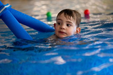 Umumi bir yüzme havuzunda mavi bir yüzme aleti kullanarak yüzmeyi öğrenen küçük bir çocuk.