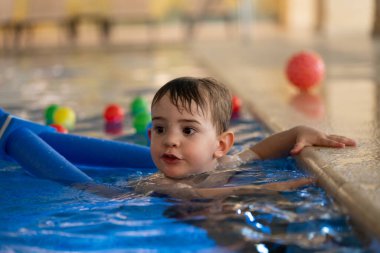 Mutlu çocuk havuzda yüzme dersleri alıyor, renkli yüzen oyuncaklarla oynuyor ve ferahlatıcı suya su sıçrıyor.