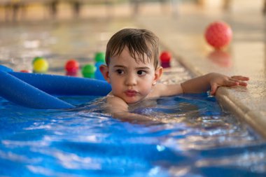 Yakışıklı erkek bebek kapalı havuzda yüzmeyi öğreniyor, kenarına tutunuyor ve yanında renkli topları olan mavi bir yüzme aleti kullanıyor.