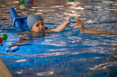 Eğitmen, yüzme şapkası takan ve yüzme havuzunda yüzme cihazı kullanan bir çocuğa pas veriyor.