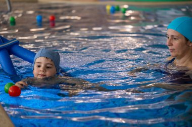 Yüzme dersi sırasında annesinin gözetiminde, renkli toplarla ve mavi bir yüzme havuzunda oynayan bir çocuk.