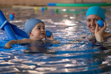 Anne ve çocuk neşeli kapalı havuz ortamında eğlenceli yüzme dersleri sırasında renkli toplarla eğleniyorlar.
