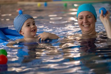 Anne ve oğul birlikte vakit geçiriyorlar, yüzme havuzunda renkli toplarla oynuyorlar, su güvenliğini ve aile bağlarını destekliyorlar.
