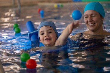 Kapalı havuzda yüzme dersi sırasında hocasına renkli bir top atan mutlu çocuk.