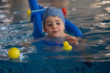 Mavi yüzme şapkası takan küçük çocuk, kapalı havuzda sarı lastik ördeklerle oynamayı, su güvenliğini ve erken çocukluk gelişimini teşvik etmeyi seviyor.