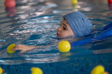 Mavi yüzme şapkası takan küçük çocuk, kapalı havuzda yüzmeyi öğrenirken renkli toplarla oynamaktan zevk alıyor.