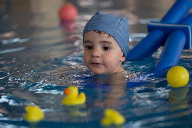 Mavi yüzme şapkası takan küçük bir çocuk, kapalı bir havuzda lastik ördekler ve diğer yüzen oyuncaklarla oynamaktan zevk alır.