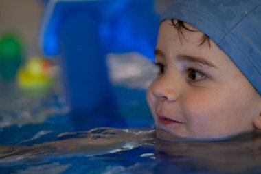 Küçük bir çocuğun havuzda oyun oynamaktan hoşlandığı, mavi bir yüzme şapkası taktığı, neşeli bir ifadeyle.