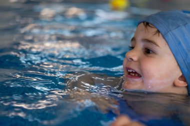 Mavi yüzme şapkası takan mutlu çocuk kapalı havuzda yüzmeyi öğreniyor, eğleniyor ve su sporlarının keyfini çıkarıyor.