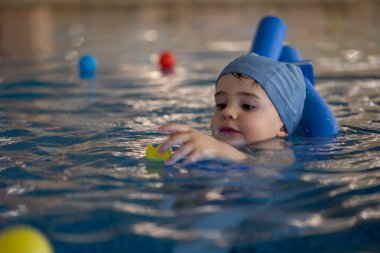 Mavi yüzme şapkası takan ve yüzme cihazı kullanan küçük bir çocuk havuzda yüzmeyi öğrenirken sarı oyuncak ördeğe uzanır.