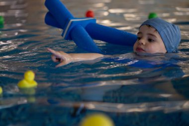 Küçük çocuk mavi yüzme cihazıyla yüzmeyi öğrenirken ve kapalı havuzda lastik ördeklerle oynarken işaret ediyor.