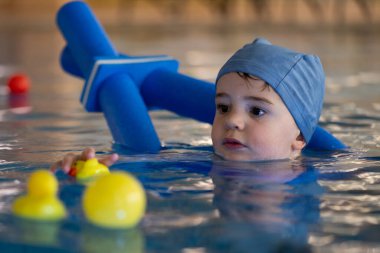 Yüzme şapkası takan ve yüzme aleti kullanan küçük bir çocuk yüzme dersi sırasında lastik ördekler ile oynar.