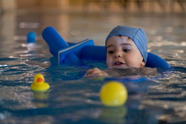 Mavi yüzme şapkası takan küçük bir çocuk, yüzme cihazıyla yüzmeyi öğrenirken sarı ördek ve topla oynamaktan zevk alır.