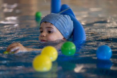 Yüzme şapkası takan ve yüzme cihazı kullanan, renkli toplarla oynayan ve kapalı havuzda yüzmeyi öğrenen bir çocuk.