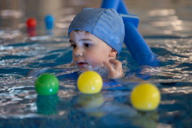 Yüzme şapkası takan ve yüzme dersi alan küçük bir çocuk kapalı havuzda renkli toplarla oynamayı seviyor.