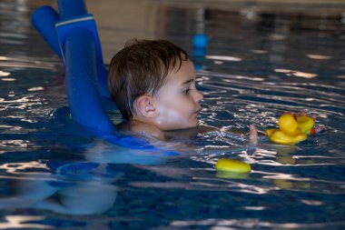 Kapalı havuzda yüzmeyi öğrenirken uçan aletler kullanan ve plastik ördeklerle oynayan küçük bir çocuk.