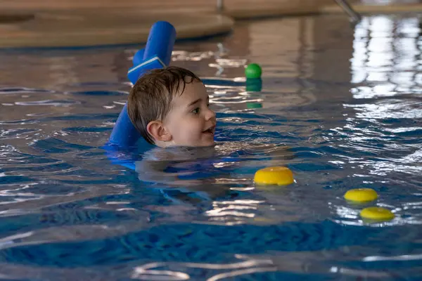 Mutlu çocuk mavi bir yüzme cihazı ve yüzme havuzunda renkli oyuncaklarla yüzmeyi öğreniyor.