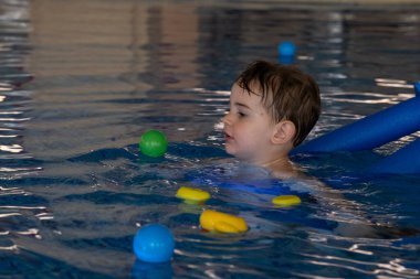 Küçük çocuk yüzme dersini seviyor, şamandıra kullanıyor, renkli toplarla oynuyor ve havuzda yüzen oyuncaklarla oynuyor.