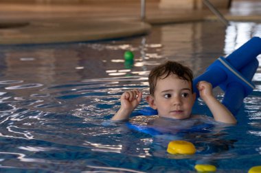 Küçük çocuk yüzme derslerini seviyor, kapalı havuzda renkli yüzme araçları kullanarak, su güvenliğini ve eğlenceyi destekliyor.
