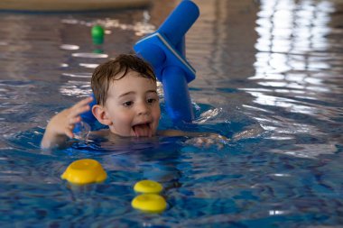 Küçük çocuk yüzme derslerini seviyor, yüzen oyuncaklarla oynarken kapalı bir havuzda yüzme aygıtı kullanıyor.